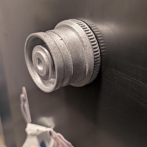 Pode incluir: Um rolha de garrafa de metal prateado com uma superfície texturizada e um anel elevado ao redor da parte superior.