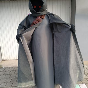 May include: A dark gray hooded cloak worn over a solid gray floor-length tunic. The cloak features a brown leather bandolier with small pouches across the chest. The hood obscures the face, revealing two glowing red circular eyes. Black gloves are worn.