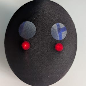 May include: A smooth, black, oval-shaped object featuring two circular, light-blue patterned patches positioned above two small, glowing red circular lights. The surface has a subtle sheen, and the overall form is rounded and symmetrical.