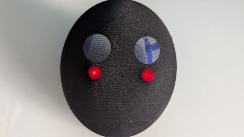 May include: A smooth, black, oval-shaped object featuring two circular, light-blue patterned patches positioned above two small, glowing red circular lights. The surface has a subtle sheen, and the overall form is rounded and symmetrical.