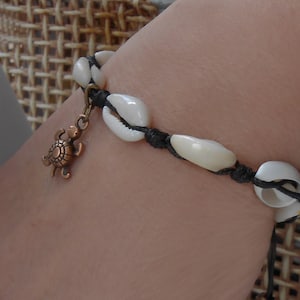 Può includere: Un braccialetto in corda nera e conchiglie bianche con un piccolo ciondolo a forma di tartaruga. Il braccialetto è regolabile con un perno di legno all'estremità.