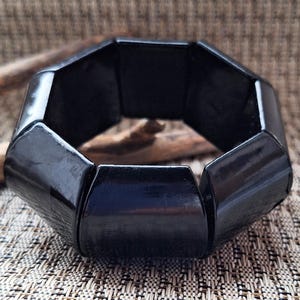 Könnte beinhalten: Ein schwarzes, geometrisches Armband mit glänzendem Finish. Das Armband besteht aus mehreren verbundenen, achteckigen Segmenten. Der Hintergrund zeigt eine strukturierte, neutral getönte Oberfläche und ein Stück Treibholz.