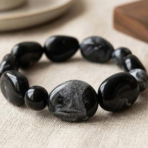 Könnte beinhalten: Ein elastisches Armband aus glatten, polierten, kieselsteinförmigen Perlen in verschiedenen Größen. Die Perlen haben eine glänzend schwarze Oberfläche, wobei einige marmorierte, grau-weiße Wirbelmuster auf ihren abgerundeten Oberflächen aufweisen.