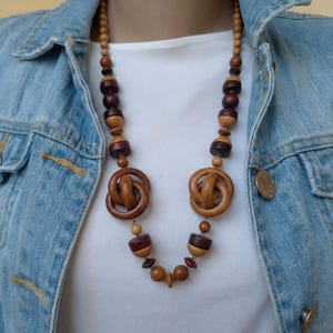 Peut inclure: Un long collier composé de perles en bois alternant des tons marron clair et foncé, de formes sphériques et cylindriques, avec deux grands pendentifs circulaires en bois noué disposés symétriquement sur la chaîne.