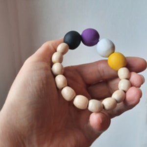 Könnte beinhalten: Ein Armband aus Holzperlen mit einer schwarzen, lila, weißen und gelben Perle. Das Armband befindet sich auf der Hand einer Person.