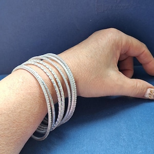 May include: A set of five silver-toned metal bangles worn on the wrist. Each bangle features a textured, grid-like pattern across its surface, creating a uniform appearance. The bracelets are thin, circular, and stacked together to form a single unit.