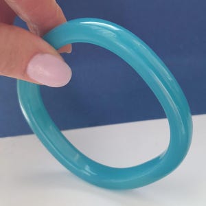 Op de afbeelding: Een doorschijnende, turquoisekleurige armband. De gladde, afgeronde armband wordt tegen een witte en blauwe achtergrond gehouden. De armband heeft een solide, cirkelvormige vorm en lijkt van glanzend materiaal te zijn gemaakt.