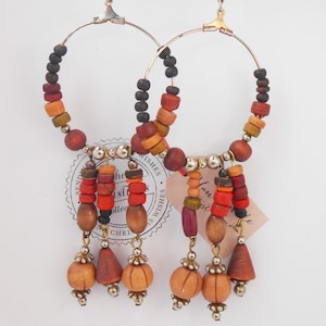 Puede incluir: Un par de pendientes de aro con cuentas de madera en tonos marrones, naranjas, rojos y negros. Los pendientes tienen un dije colgante con cuentas de madera y un acento de metal plateado.