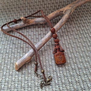 Pode incluir: Um colar de couro castanho com um pingente de madeira esculpida em forma de garrafa de vinho. O pingente apresenta um desenho de uvas. O colar tem um cordão castanho e contas de madeira. O fecho é de cor bronze.