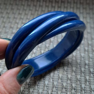 Könnte beinhalten: Ein glänzendes, marineblaues Armreif-Armband mit einem verdrehten Design. Das Armband scheint aus einem festen Material zu bestehen und hat eine glatte, reflektierende Oberfläche. Ein klassisches Schmuckaccessoire.