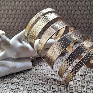 Puede incluir: Un brazalete dorado con textura martillada, que se enrolla en espiral alrededor del brazo. El brazalete se exhibe junto a una escultura blanca de una figura reclinada. El fondo es una superficie texturizada de tonos neutros.