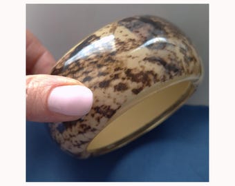 Peut inclure: Un large bracelet jonc ovale avec un motif tacheté dans les tons marron et crème. L'intérieur du bracelet est de couleur crème unie. Le bracelet est tenu par une main avec un ongle peint en rose.