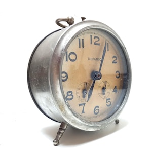 May include: A silver alarm clock with a white face and black numbers. The clock has a small second hand and a larger minute hand. The clock is labeled "Dinamic".