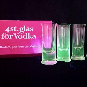 Könnte beinhalten: Vier klare Schnapsgläser aus Glas stehen neben einer rechteckigen pinkfarbenen Schachtel. Die Schachtel trägt den weißen Schriftzug 4st.glas för Vodka und Boda/Signe Persson-Melin. Die Gläser haben eine zylindrische Form mit leicht ausgestelltem Rand.
