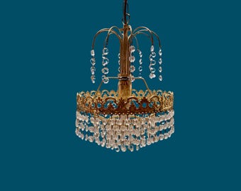 May include: A vintage gold-toned chandelier with cascading crystal droplets. The chandelier features a decorative filigree design around the base and a central column. The background is a teal color.