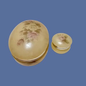 Puede incluir: Dos joyeros decorativos, uno en forma de huevo y otro redondo. Ambos son de color crema con un diseño floral de rosas rosas y verdes. Los joyeros tienen una superficie reflectante.