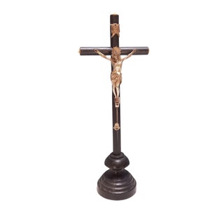 May include: A dark brown wooden crucifix with a gold-coloured Jesus figure. The cross has a decorative base and gold accents. The figure of Jesus is detailed, with arms outstretched. The crucifix is designed to stand upright.