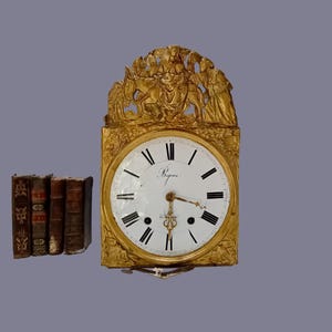 Può includere: Un orologio antico color oro con quadrante bianco e numeri romani neri. L'orologio ha una parte superiore ornata con figure ed è accanto a una pila di vecchi libri con copertine in pelle marrone.