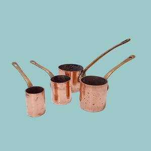 May include: Four copper saucepans with brass handles are arranged against a teal background. The pans vary in size, with a shiny, reddish-brown exterior and a darker interior. The handles are long and curved, adding to the vintage aesthetic.