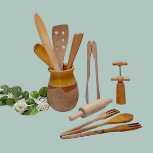 Könnte beinhalten: Eine Sammlung von Küchenutensilien aus Holz, darunter Löffel, Spatel und Zangen, die in einem rustikalen Keramiktopf präsentiert werden. Ein Nudelholz, ein Korkenzieher und Gabeln sind ebenfalls vorhanden. Auf hellgrünem Hintergrund.