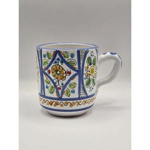 May include: A white ceramic mug featuring hand-painted blue, yellow, and green floral and geometric patterns. The design includes diamond shapes, circular motifs, and a horizontal band of orange dots near the base, with a curved handle on the side.