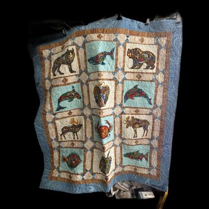 May include: A handmade quilt featuring a patchwork design with various animal illustrations. The quilt showcases a wolf, fish, bear, dolphin, eagle, moose, crab, and other creatures. The quilt has a light blue border and a beige and blue color scheme.