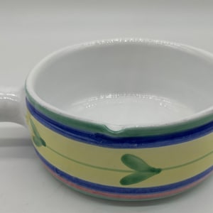 May include: White ceramic soup bowl with a handle. The bowl has a yellow band with green leaf designs, bordered by blue, green, and pink stripes. The interior is white.