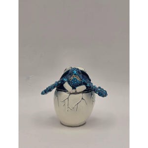 Può includere: Un portagioie decorativo a forma di uovo argentato con un design a guscio incrinato. Dalla parte superiore emerge una statuetta di tartaruga marina blu con una superficie testurizzata e scintillante. La tartaruga ha le pinne spiegate.