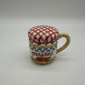 Può includere: Una tazza in ceramica con coperchio, con un motivo a scacchi rosso e bianco sul coperchio. La tazza ha un manico ed è decorata con la parola "preserves" e illustrazioni di mele. La tazza ha un motivo a scacchi blu e bianco al centro.