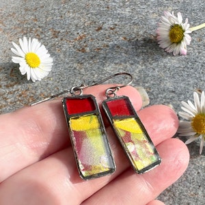 May include: A pair of rectangular drop earrings featuring a silver-toned metal frame. Each earring contains a solid red glass section at the top and a larger lower section filled with a marbled blend of yellow, red, and green colours.