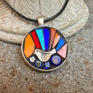 May include: A round pendant necklace featuring a silver-toned bird charm with a spiral detail. The pendant is filled with a mosaic of colourful glass shards in orange, red, light blue, dark blue, and peach, accented by small floral millefiori beads.