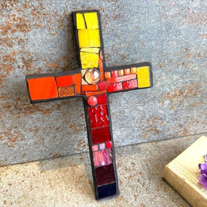 May include: A mosaic cross featuring a gradient of colours ranging from bright yellow at the top to deep red and dark purple at the bottom. The cross is constructed from various small, angular glass tiles and includes a single clear circular glass bead.
