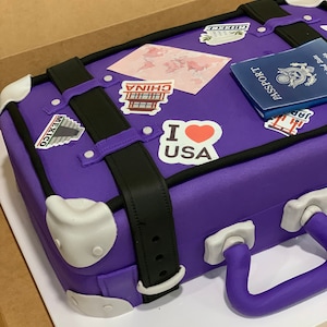 May include: A purple suitcase cake decorated with travel stickers, including a map, "I love USA", "China", "Mexico", and a passport.