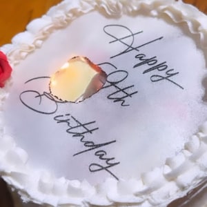 Puede incluir: Pastel blanco con glaseado blanco y un borde rizado. El pastel está decorado con un mensaje impreso que dice "Feliz cumpleaños número 50". Hay una vela encendida en el centro del pastel.