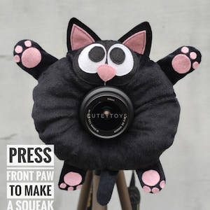 May include: Black cat camera lens cover with pink paws and ears. The cover is on a tripod and says "PRESS FRONT PAW TO MAKE A SQUEAK NOISE" on the bottom.