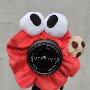 May include: A red plush camera cover with large white eyes and a small cookie attached. The camera lens is visible, with the text "UTEYTOYS" and "Nikon" printed on it. The camera is mounted on a tripod.
