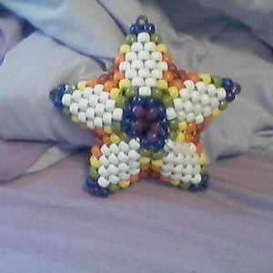 May include: A beaded star in a five-point design. The star is made of small, colorful beads in white, orange, yellow, green, blue, and purple. The star is resting on a purple surface.