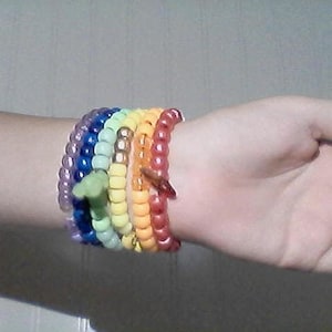 Könnte beinhalten: Eine Sammlung farbenfroher Perlenarmbänder in Regenbogenfarben, darunter Lila, Blau, Grün, Gelb, Orange und Rot. Die Armbänder werden am Handgelenk getragen und zeigen die lebendigen Farben und das verspielte Design. Die Perlen scheinen aus Kunststoff zu sein.