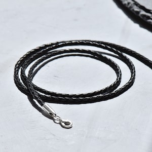 Può includere: Una collana in cordoncino di cuoio nero con una chiusura argentata. Il cordoncino è intrecciato e ha una texture liscia.
