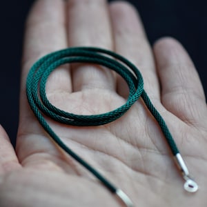 May include: A coiled, dark green cord necklace with silver-coloured metal ends and a clasp. The cord is tightly woven, and the necklace is displayed against a blurred background.
