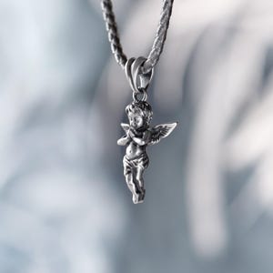 Romantic silver angel pendant hanging from a textured braided cord, casting a soft, ethereal glow against a dreamy, blurred background