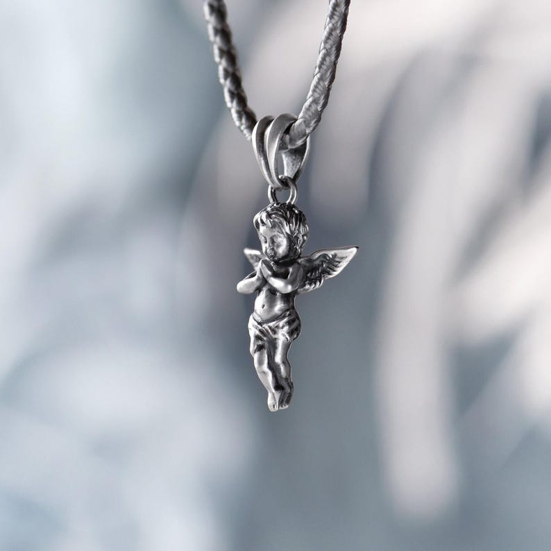 Romantic silver angel pendant hanging from a textured braided cord, casting a soft, ethereal glow against a dreamy, blurred background
