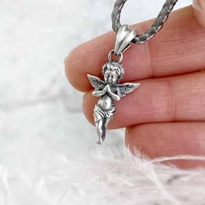 Close-up of a detailed silver angel pendant with tiny wings and a praying pose, held gently between fingers, set against a soft, white background for a serene and divine touch