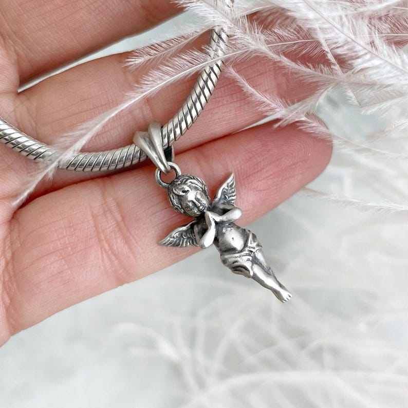 Close-up of a silver angel charm in prayer, held tenderly in hand, framed by soft white feathers that highlight its serene elegance and protective symbolism