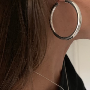 May include: A large, polished silver hoop earring with a thick, rounded profile. The earring is worn in the earlobe. Additionally, a thin silver chain necklace with a small, smooth, oval-shaped pendant rests against the neck area.