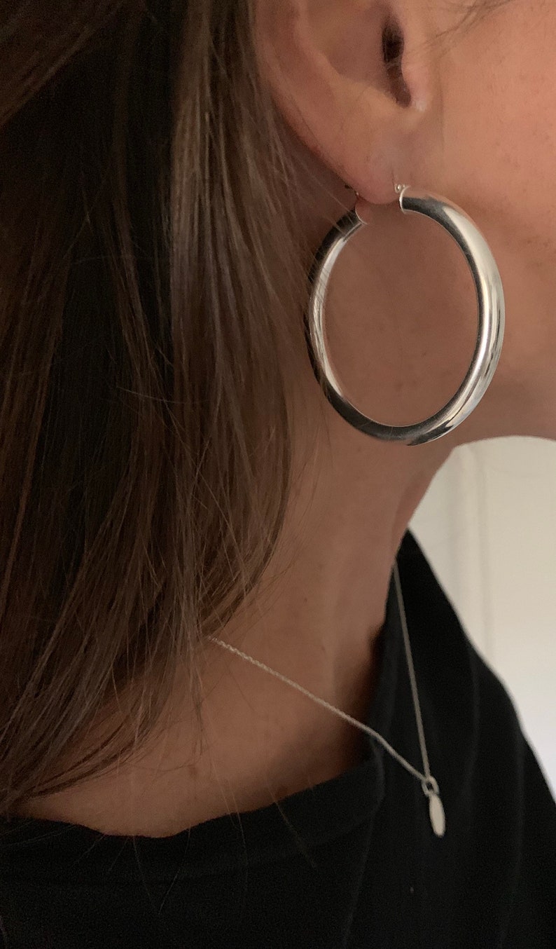 May include: A large, polished silver hoop earring with a thick, rounded profile. The earring is worn in the earlobe. Additionally, a thin silver chain necklace with a small, smooth, oval-shaped pendant rests against the neck area.