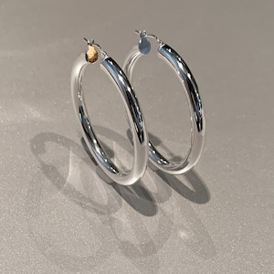 May include: A pair of polished silver-tone hoop earrings with a smooth, rounded tubular design. Each earring features a hinged post closure at the top, showcasing a reflective metallic surface that catches the light with a clean, circular silhouette.