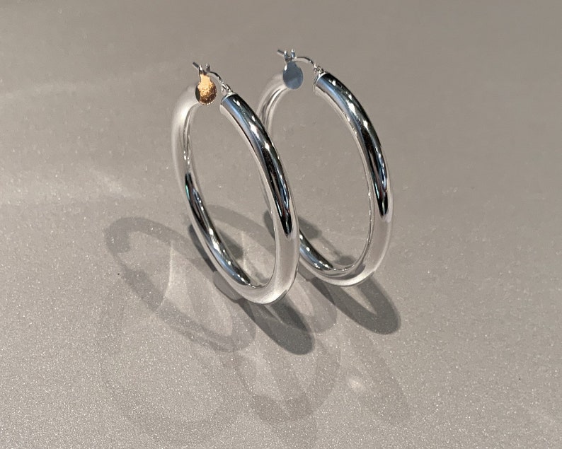 May include: A pair of polished silver-tone hoop earrings with a smooth, rounded tubular design. Each earring features a hinged post closure at the top, showcasing a reflective metallic surface that catches the light with a clean, circular silhouette.