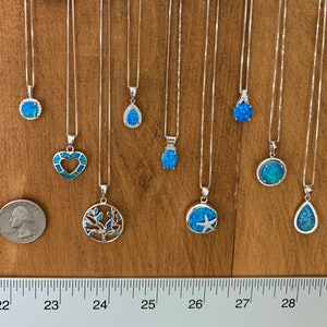 May include: Ten silver necklaces with blue opal pendants in various shapes, including a heart, a tree, a starfish, and a teardrop. The necklaces are displayed on a wooden surface with a ruler for size reference.