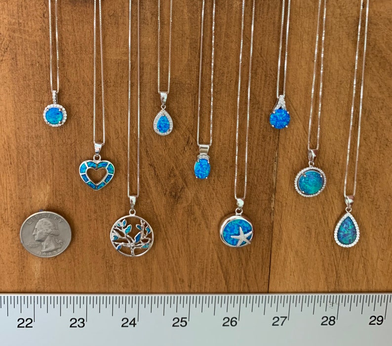 May include: Ten silver necklaces with blue opal pendants in various shapes, including a heart, a tree, a starfish, and a teardrop. The necklaces are displayed on a wooden surface with a ruler for size reference.